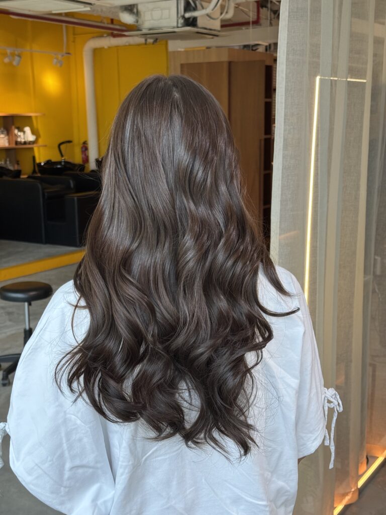 Long ash brown hair color on naturally dark hair with a soft root gradient and curled finish at a Japanese hair salon in Singapore.